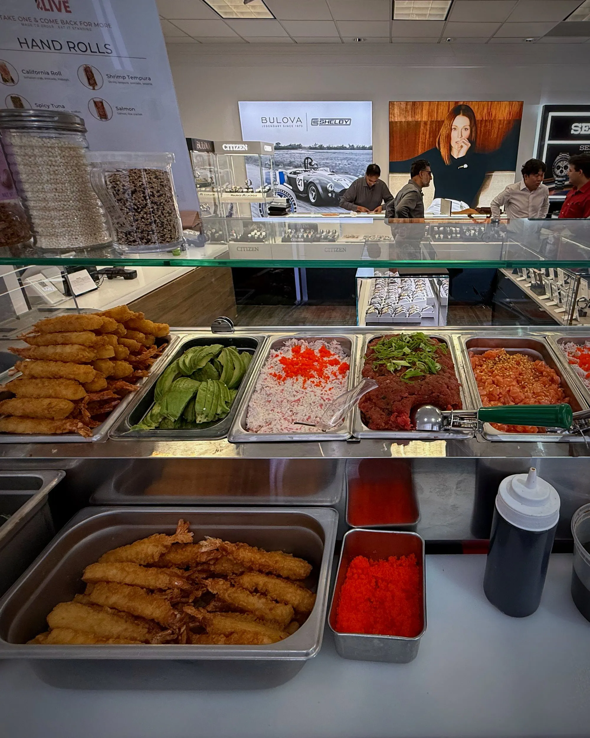 The counter mid-service — salmon, tuna, crab, avocado and tempura in their trays under glass, guests behind